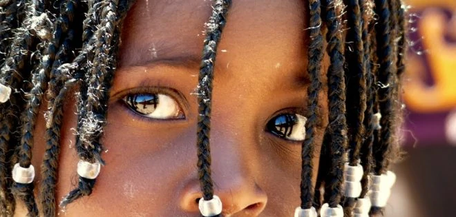 A child with decorated braids looks thoughtfully into the camera.