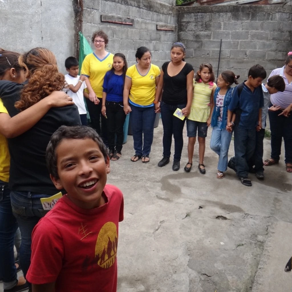 Children and young people stand in a circle in the backyard of a house and hold each other's hands. Some of them have put on blue and yellow T-shirts.
