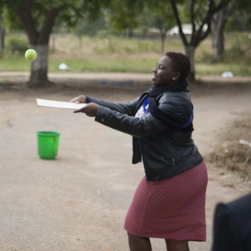 Two women blanching a tennis ball with a piece of cardboard in their hands. They throw the tennis ball to each other with the cardboard.