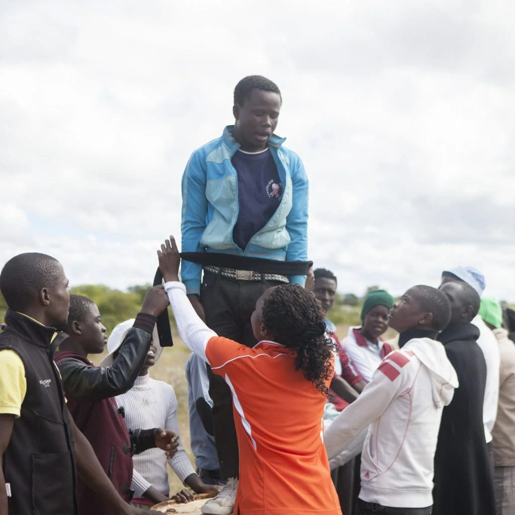 A black youth stands on a pedestal and is tied up with a black ribbon. Other youths stand around him for his protection.