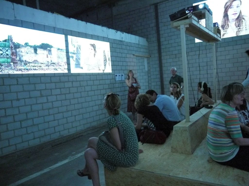 Exhibition visitors sit with headphones in a darkened room with various projectors.