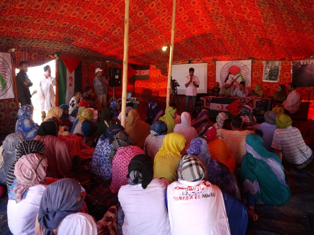 Dozens of people sit on the ground in a desert tent and watch an event.