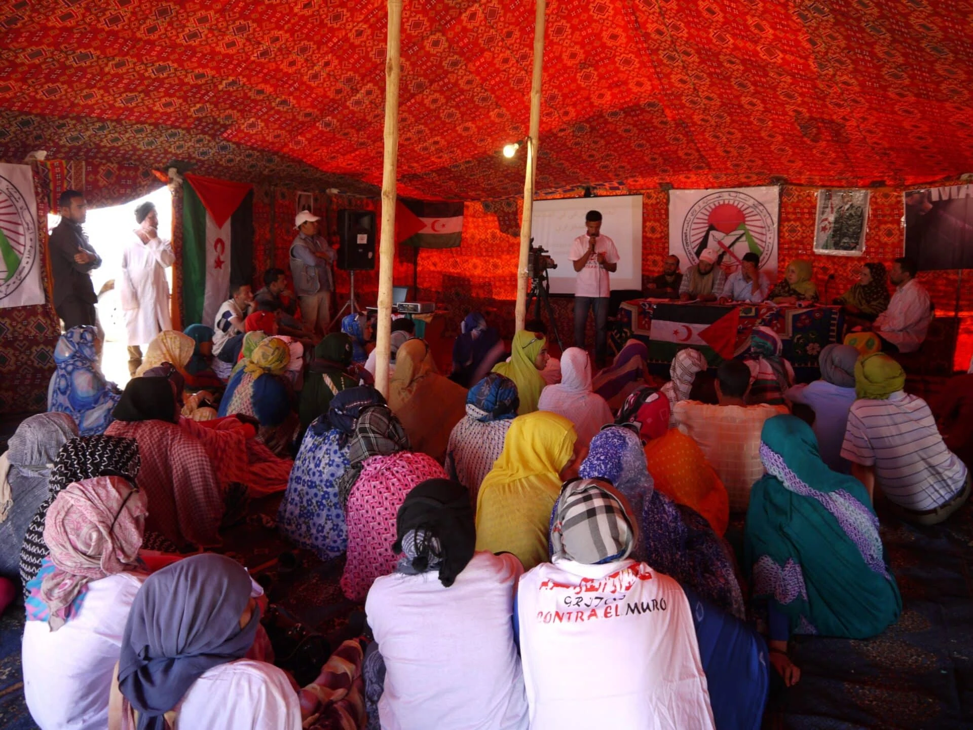 Dutzende Menschen sitzen im einem Wüstenzelt am Boden und verfolgen eine Veranstaltung.