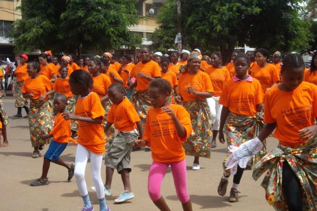 Women and girls in orange T-shirts and uniform skirts dance uniformly in a public square.