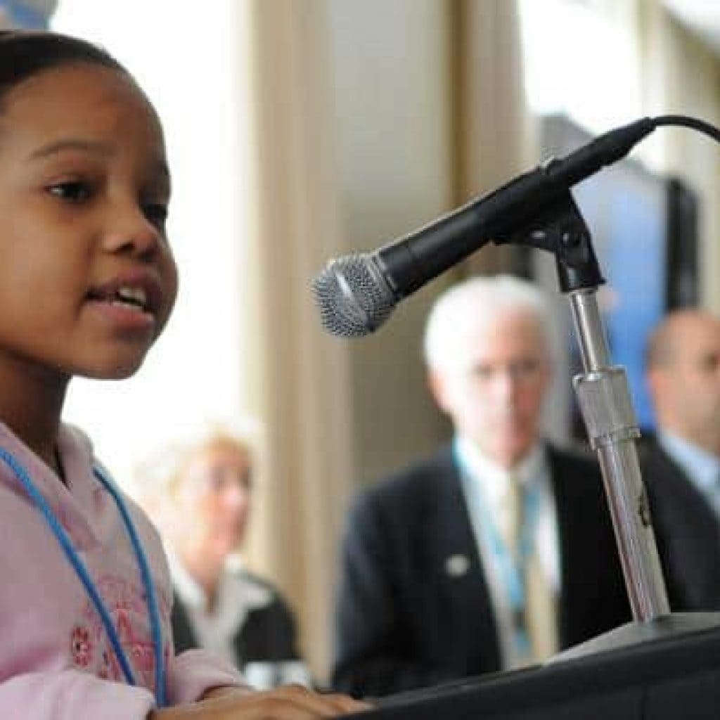 Schwarzes Mädchen spricht in ein Mikrofon auf einem Stehpult.