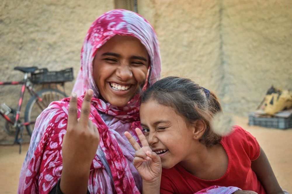 Zwei lachende Mädchen, eine mit Kopftuch. Sie machen das Peace-Zeichen.