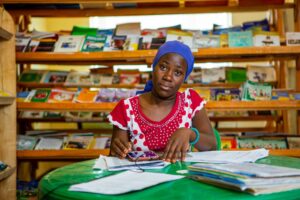 Aronia sits at a green plastic table in front of a pile of papers. In the background wooden shelves with magazines.