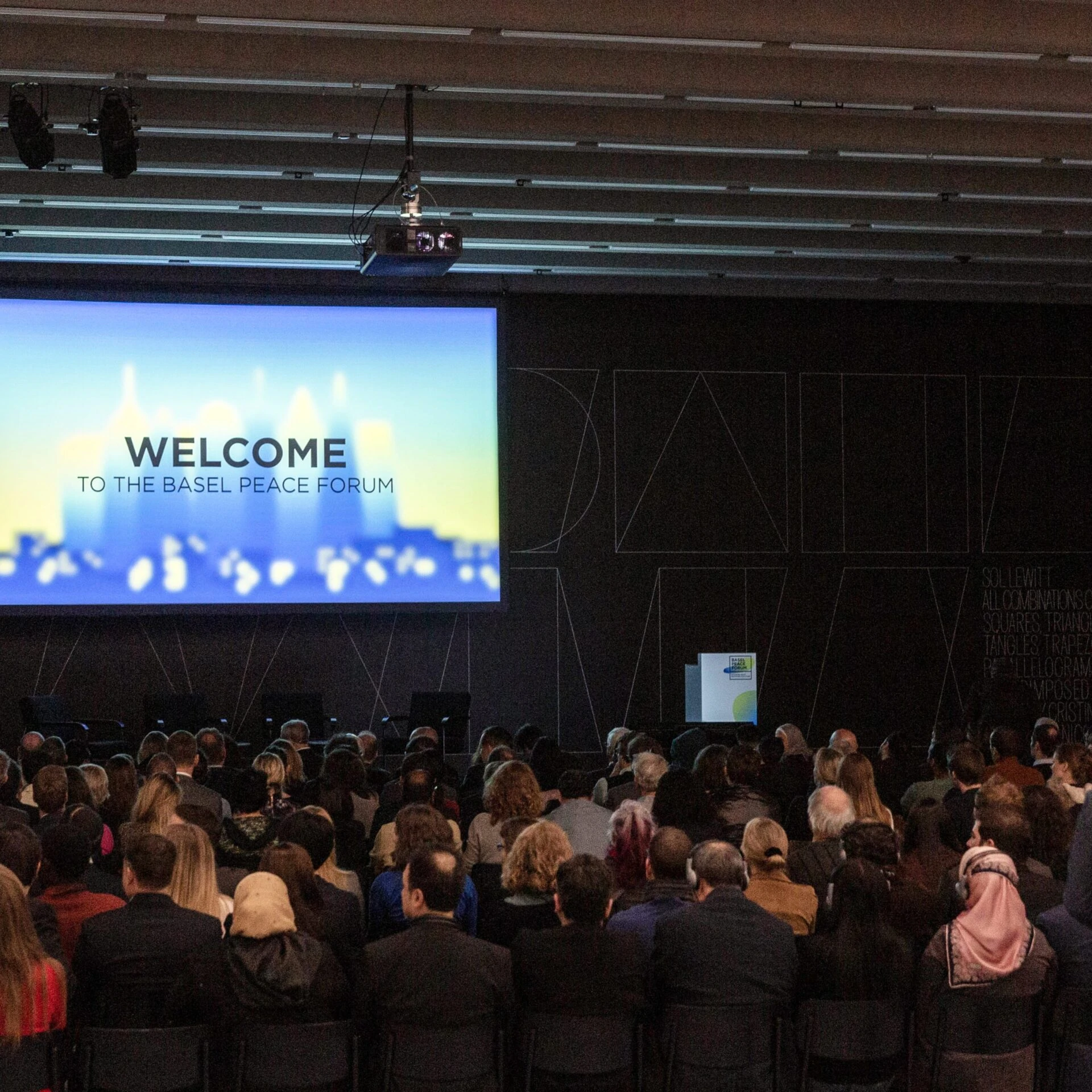 Leinwand mit Beamerbild: Welcome to the Basel Peace Forum. Publikum im abgedunkelten Saal.