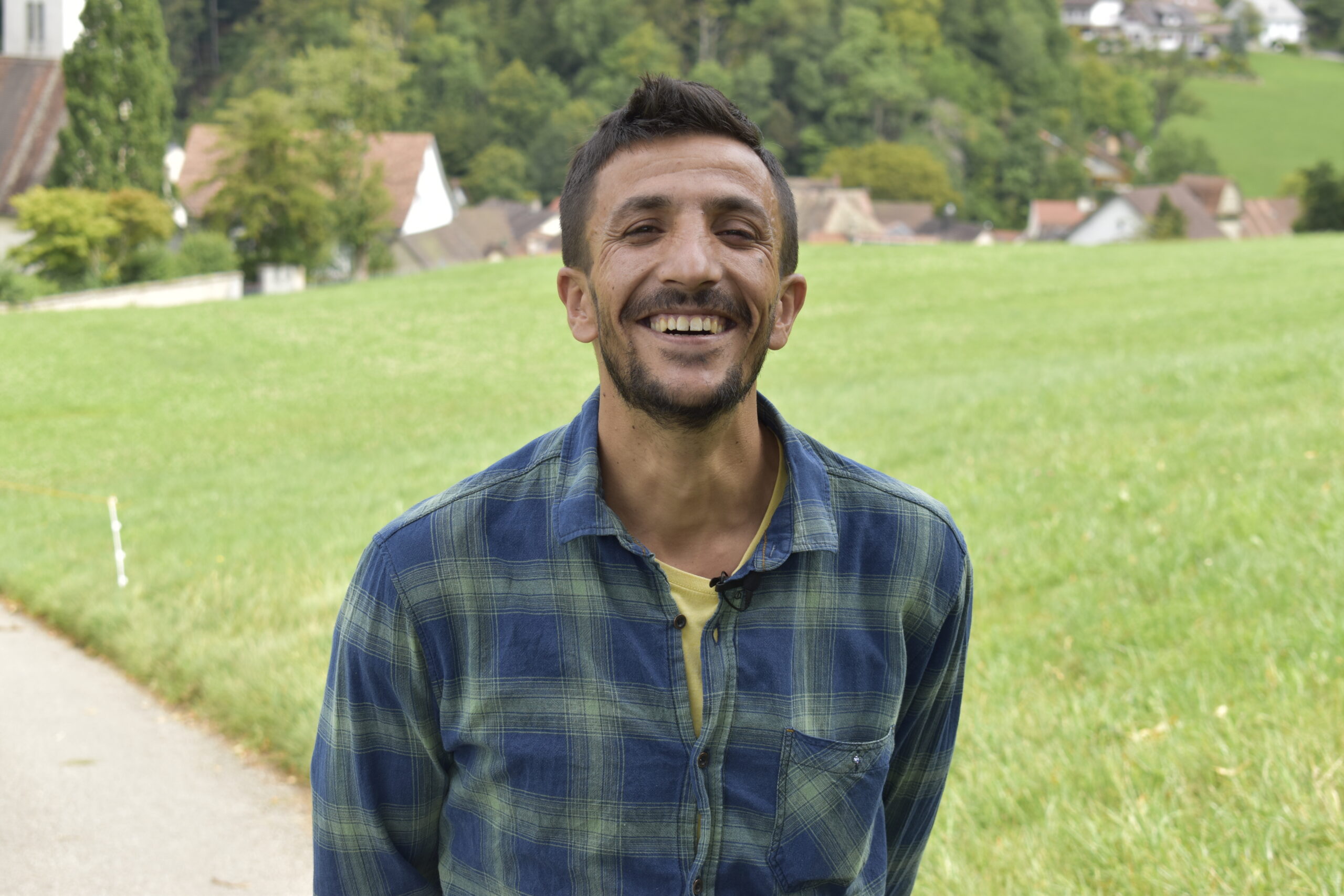 A young man, in a checkered shirt, laughs into the camera, a green meadow behind.