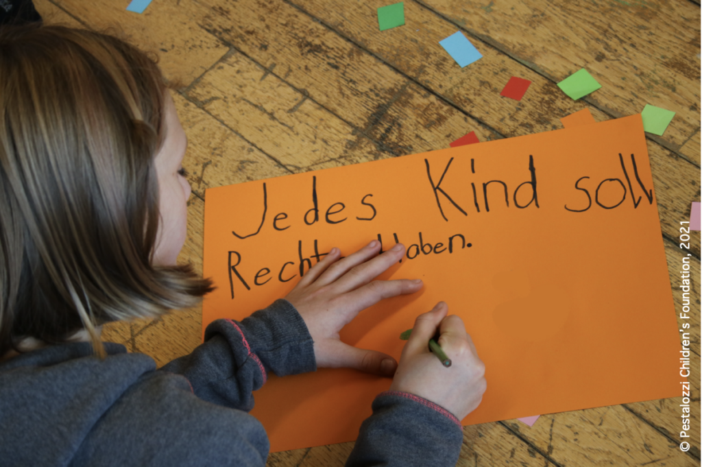Ein-Kind-im-Schulalter-schreibt-auf-einem-grossen-Papier-sitzend-auf-dem-Boden-Punkt-Jedes-Kind-soll-Rechte-haben.