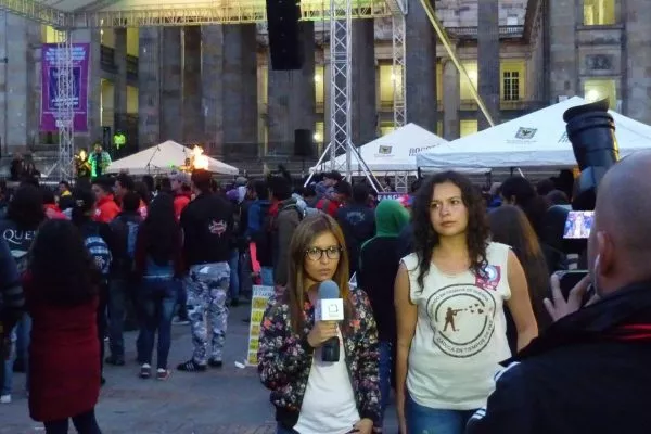 Eine junge Frau von ACOOC wird im Juli 2014 an einem Anti-Militär-Festival in Bogota vom lokalen Fernsehsender Canal Capital interviewt. Im Hintergrund ist eine Bühne zu sehen.