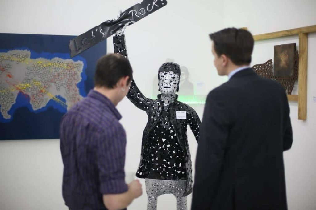 Zwei Besucher betrachten eine lebensgrosse Figur aus Metall mit einem Schild in der Hand. Text: Let's Rock!