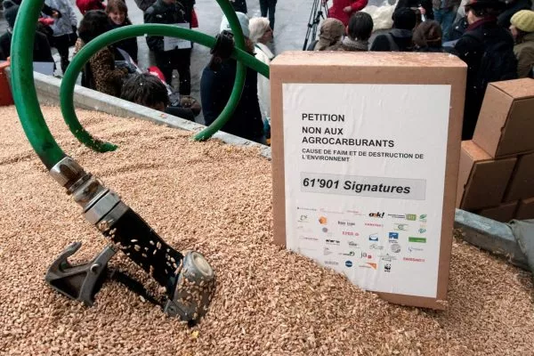 Im Hintergrund die Vertreter der Plattform Agrotreibstoffe und im Vordergrund ein Benzinzapfschlauch der in einem Haufen Getreide steckt.